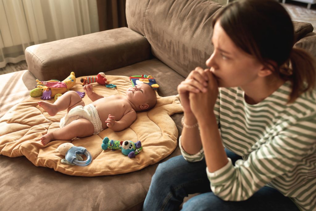 Worried mother sitting on sofa while her baby lies on a play mat with toys