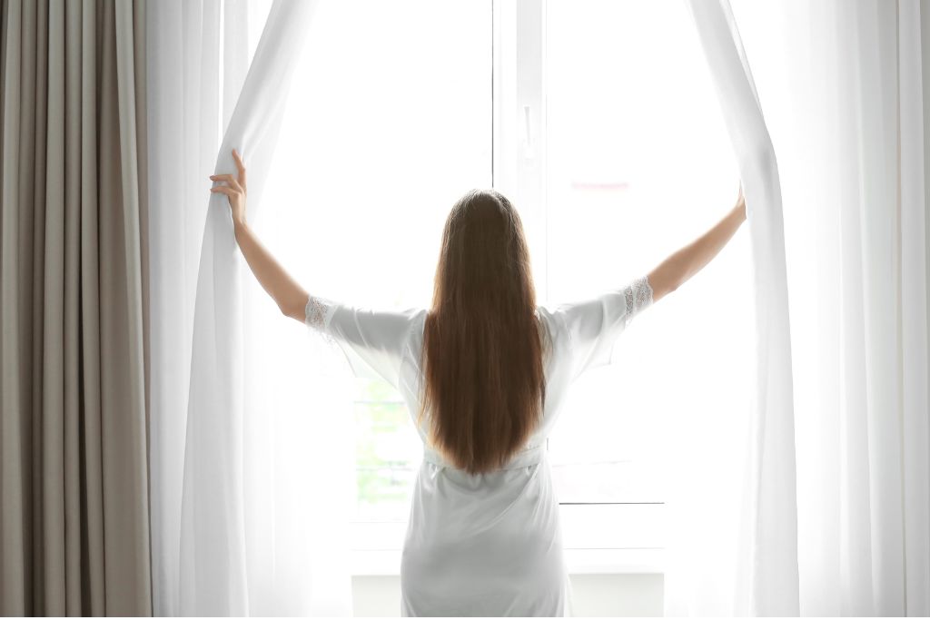 Woman opening white curtains with sunlight streaming in, symbolising hope and recovery