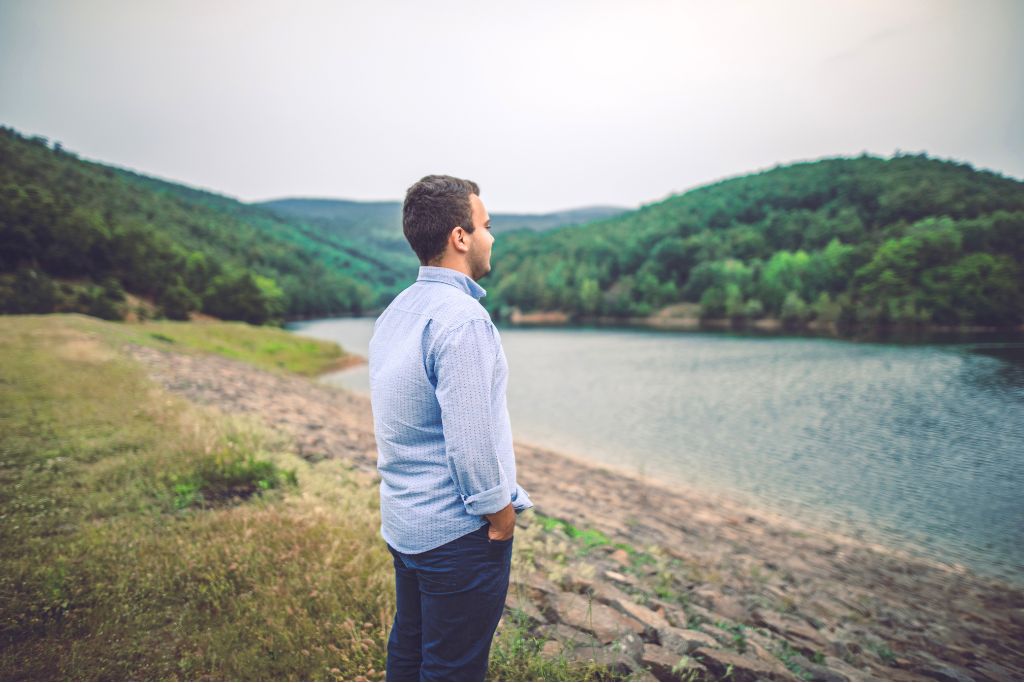 Man standing alone by a peaceful lake, looking out at the water and surrounding trees