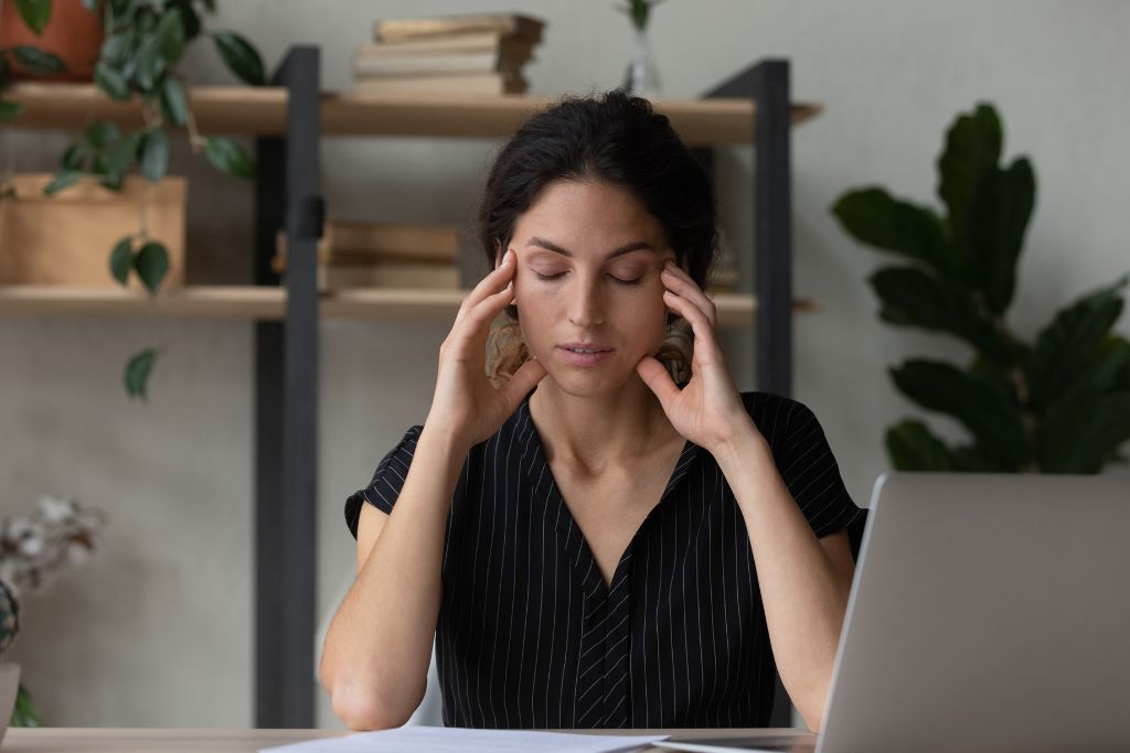 A woman with eyes closed and hands at her temples, taking a quiet moment at her desk