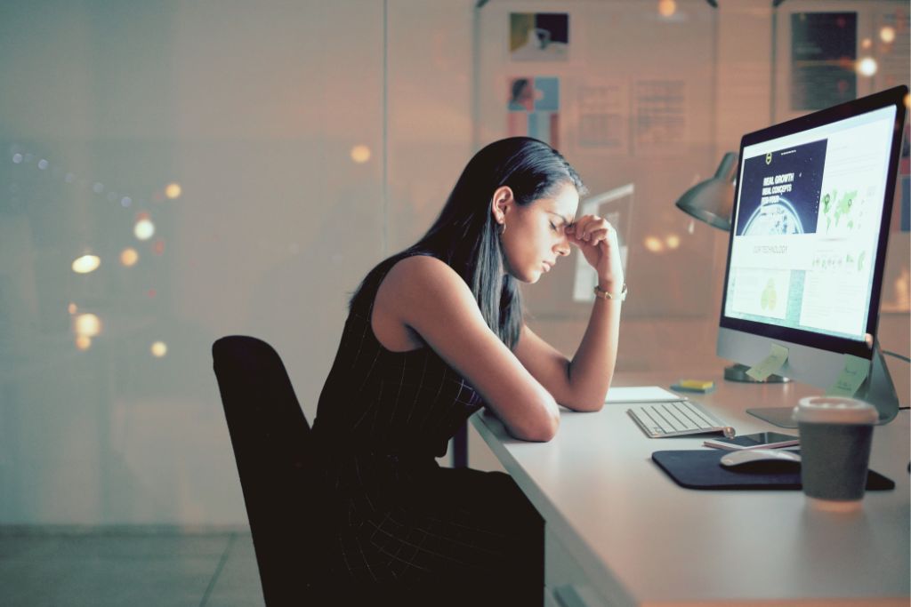 Woman sitting at desk with head resting on hand, looking tired and reflective in front of computer screen