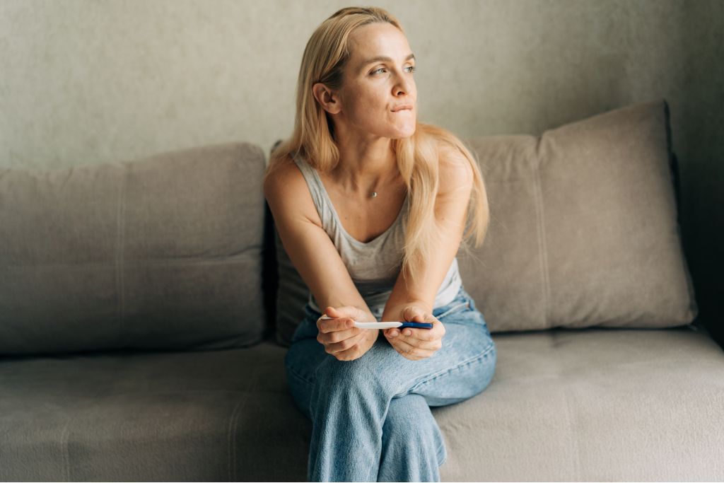 Woman sitting on sofa holding a pregnancy test and looking uncertain