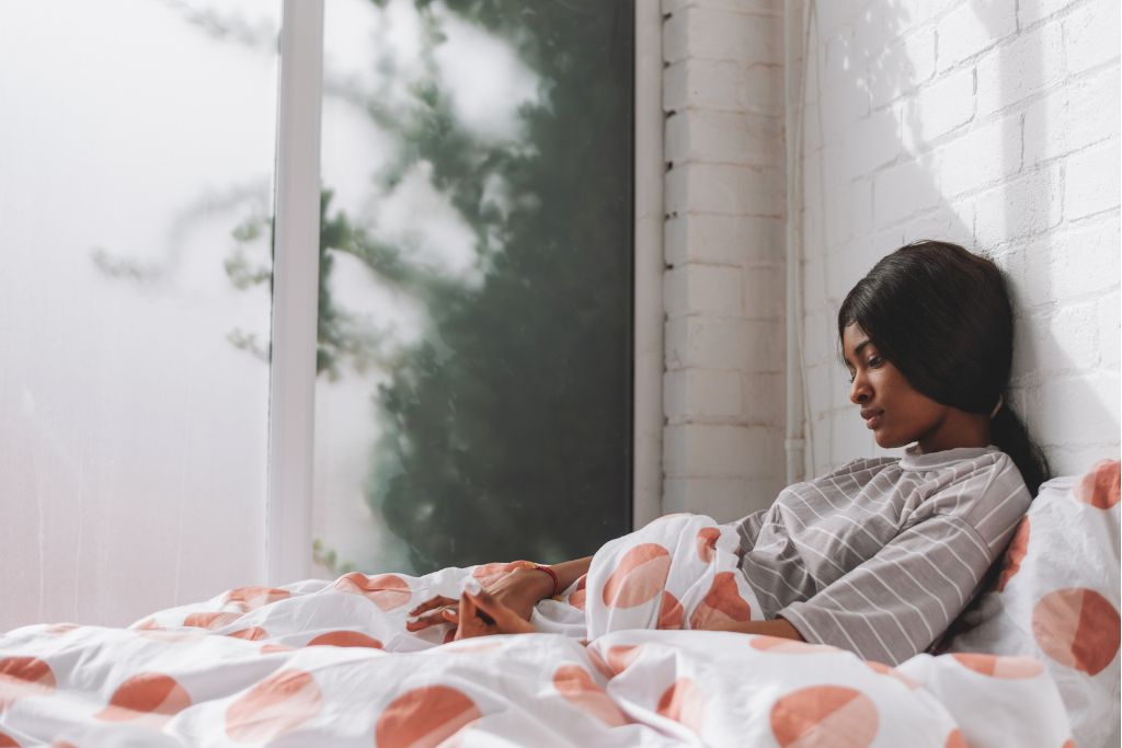 Woman sitting quietly in bed in soft morning light, looking down in thought.