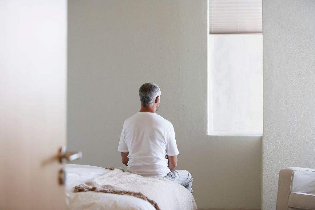 Man sitting alone on a bed facing a window, suggesting avoidance or emotional withdrawal