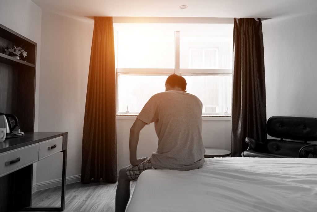 Man sitting at the edge of a bed in the morning light, appearing tired and reflective