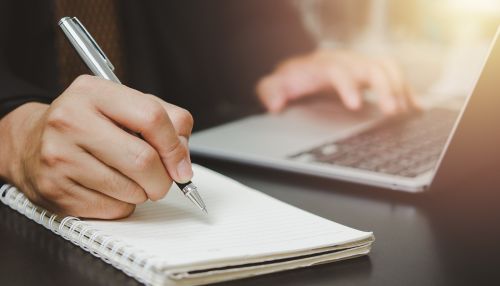 Close-up of a person writing in a notebook beside a laptop, representing evidence-based therapeutic practice