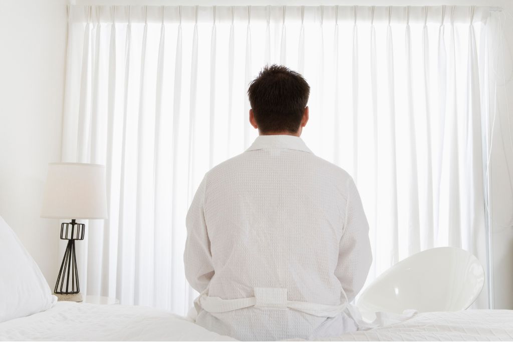 Man sitting on a bed facing drawn curtains, suggesting emotional distance or inner conflict